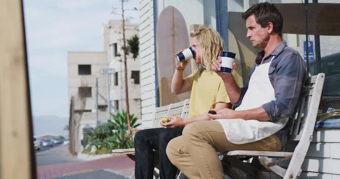Two Caucasian male surfboard makers sitting on a bench and drinking takeaway coffee