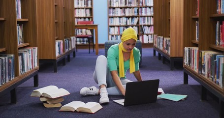 An Asian female student wearing a yellow hijab studying in a library and using laptop - Powered by Adobe