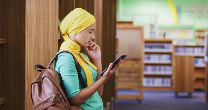Asian female student wearing a yellow hijab leaning against bookshelves and using tablet