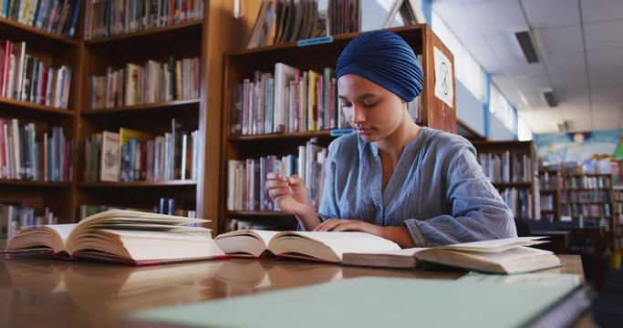 Asian female student wearing a blue hijab sitting at a desk with open books and taking notes