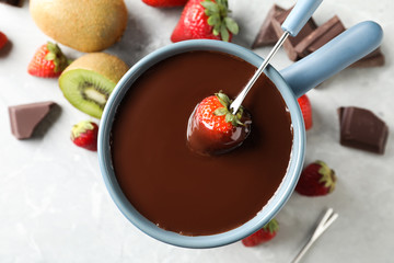 Dipping strawberry into fondue pot with chocolate on light table, top view