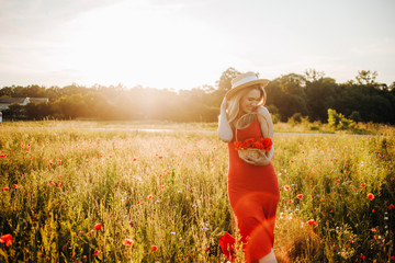 Fashionable blonde in a straw hat and red dress posing in a poppy field holding a basket with...