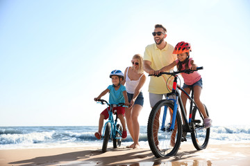 Happy parents teaching children to ride bicycles on sandy beach near sea