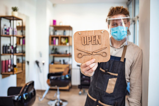 Hairdresser stands in a hairdressing studio with protective face mask, during COVID-19. Small business survival after covid-19 pandemic. Owner man in apron with medical mask, hair comb and scissors.