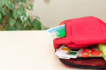 Back to school. Education and social distance concept. Protective medical mask, antibacterial hand cleaning gel (alcohol), school supplies lie next to a backpack on a light background