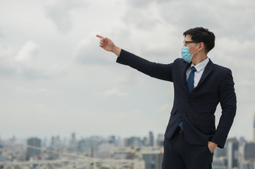 Young handsome asian businessman standing wearing mask of protect the pollution and virus and pointing the finger on building urban cities background.