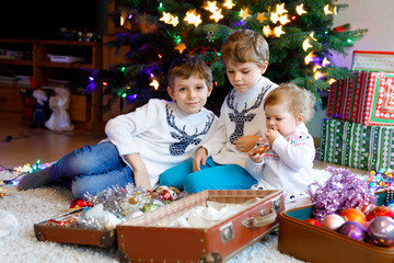 Two little kid boys and adorable baby girl decorating Christmas tree with old vintage toys and balls. Family preaparation celebration of family holiday. Three children, brothers and sister at home.