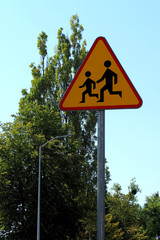 the road sign is made of metal triangular shape in orange color with the image of children against a background of green trees and blue sky . Careful children