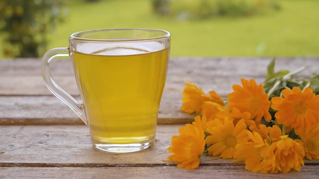 Close-up transparent cup of calendula tea on the wooden table in the garden at summer day. A bouquet of calendula flowers on the table. Herbal medicine concept. Dolly shot