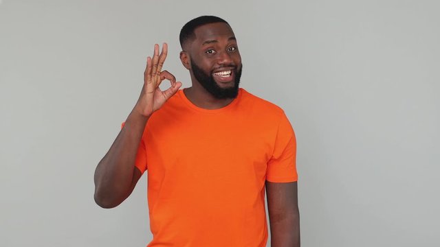 Young bearded african american guy 20s in orange basic casual t-shirt look at camera with charming smile say wow it is okay showing doing ok hand gesture isolated on grey color gray background studio