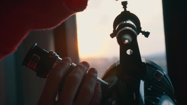Girl next to the window looking through the telescope at the sky in her bedroom at sunset. Hobbie while you have to be locked up at home. Confinement, being at home.