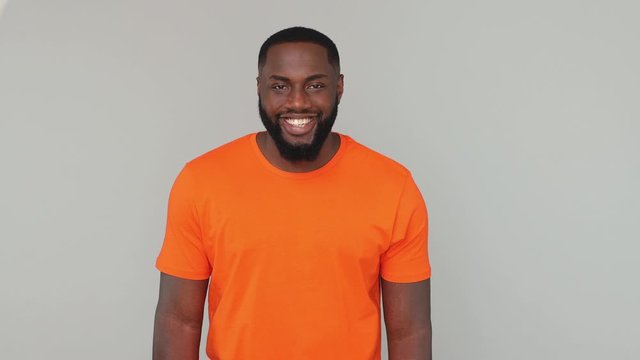 Young calm bearded african american man guy male 20s in orange basic casual t-shirt looking at camera with charming smile isolated on grey color gray background in studio