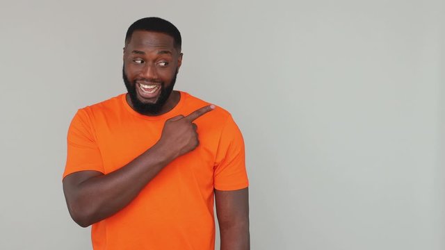 Young fun african american man in orange basic casual t-shirt look at camera pointing finger hand on empty place aside workspace mockup for your advertising isolated on gray color background in studio