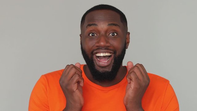 Close up young bearded african man in orange basic casual t-shirt looking at camera with open eyes shocked surprised say wow found out heard good news isolated on grey color gray background in studio