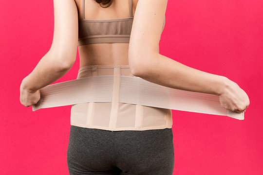 Back view of pregnant woman putting on supporting bandage to reduce backache at pink background with copy space. Cropped image of orthopedic abdominal support belt concept