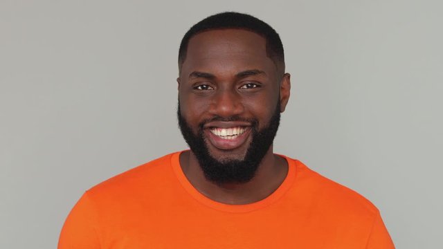 Close up young confident bearded african american man guy male 20s in orange basic casual t-shirt looking camera with charming smile blinking wink eye isolated on grey color gray background in studio