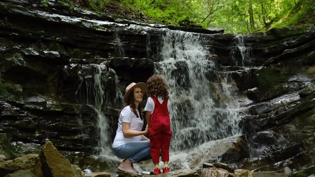 Cheerful mom in hat and cute little daughter sitting on edge of cliff near waterfall, enjoys beautiful summer views. Dreamy and inspirational family walk through. Outdoor activities travel concept