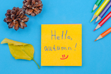 Yellow sheet of paper with the inscription Hello, autumn on a blue background with a yellow tree leaf, cones, colored pencils, copy space, top view. Greeting of the season