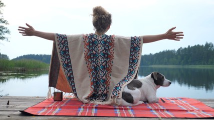 Woman in cozy poncho wide spreading her hands appreciating the nature and her time with white and brown dog on the dock of the lake at sunset. Self care concept.