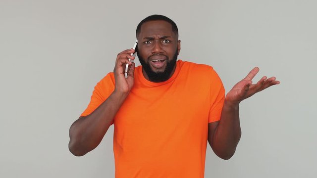 Young dissatisfied irritated confused fun african american man in orange basic casual t-shirt hold do not want talking to girlfriend using mobile phone isolated on grey color gray background in studio