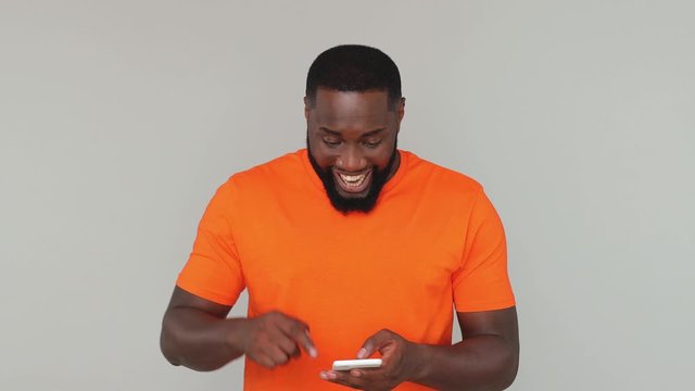 Young shocked happy win bearded african american man guy male 20s in orange basic casual t-shirt look surprised say wow hold using mobile cell phone isolated on grey color gray background in studio