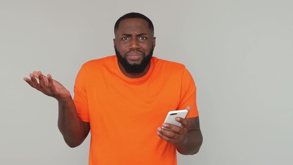 Sad shocked upset young bearded african american man 20s in orange basic casual t-shirt screaming using mobile cell phone just found out bad fake news isolated on grey color gray background in studio - Powered by Adobe