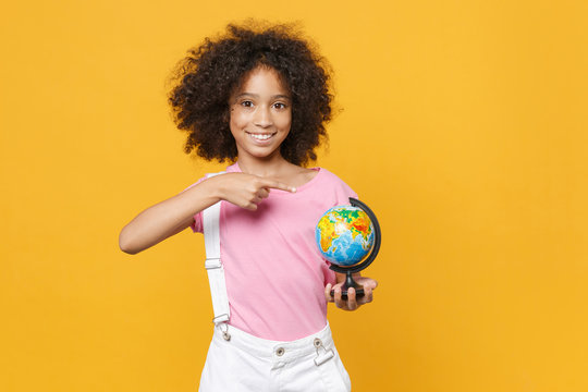 Smiling little african american kid girl 12-13 years old in pink t-shirt isolated on yellow background studio. Childhood lifestyle concept. Mock up copy space. Point index finger on Earth world globe.