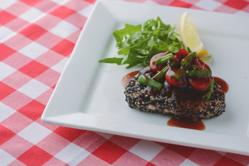 Beef stir-fry steak covered in sesame seeds with vegetables and tomato sauce served on white plate over plaid tablecloth