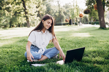 student girl with laptop outdoors. Smiling woman sitting on the grass with a computer, surfing the Internet or preparing for exams. Technology, education and remote work concept. Soft selective focus.