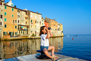 Fototapeta na wymiar Funny little girl looking through vintage binoculars on sunny summer day. Rovinj town in background. Travel and adventure concept.