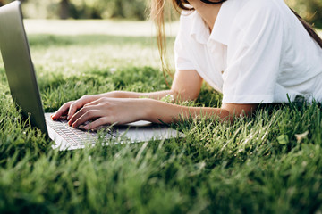 student girl with laptop outdoors. woman lying on the grass with a computer, surfing the Internet or preparing for exams. Technology, education and remote work concept. Soft selective focus.