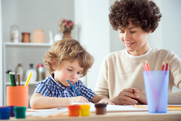 Happy mother and baby son drawing together in a room lit by the sun