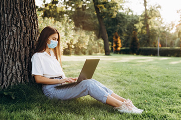 beautiful and young girl who studies and works with a laptop with a surgical mask in the park, blogger and smart worker during the epidemic, home schooling, distance learning. Soft selective focus.