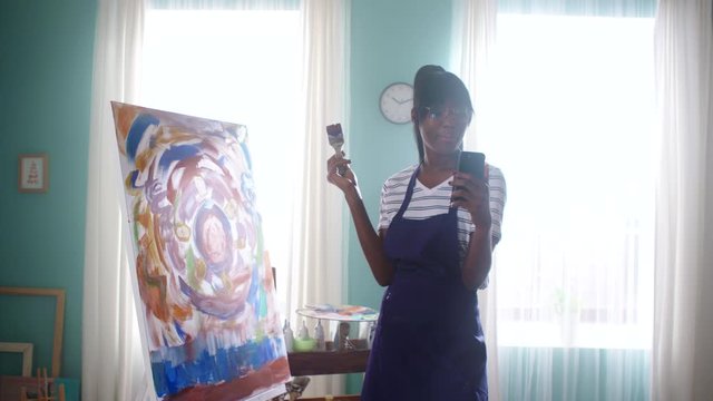 Young African woman, artist, is filming video lesson about abstract art in studio, using her mobile phone, sharing experience and knowledge, demonstrates example of latest work, Slow motion.