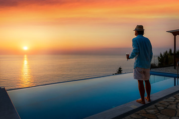 Ein attraktiver Mann mit Hut steht am Pool und genießt den Sommer Sonnenuntergang über dem Meer...
