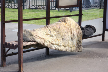 Homeless man sleeps under a blanket at a public transport stop