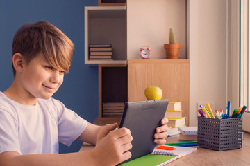 Schoolboy using his tablet during online lesson or training classes, sitting at wooden table near window. Distance education and online studying concept. Back to school, new normal