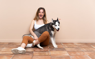 Young pretty woman with her husky dog sitting in the floor at indoors surprised and pointing finger to the side