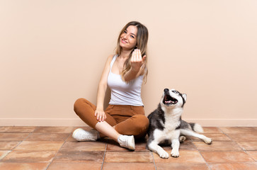 Young pretty woman with her husky dog sitting in the floor at indoors inviting to come with hand. Happy that you came