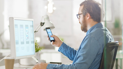 Young Focused Software Developer Compares Smartphone Application Design with Same App Design Shown on Desktop Computer Screen. He is Working in a Bright Modern Loft Office.