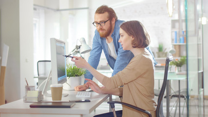 Obraz premium Młody mężczyzna menedżer i piękna kobieta omawiają kwestie związane z pracą w miejscu pracy. Ona używa komputera, a on stoi obok niej. Wskazuje na coś na ekranie.