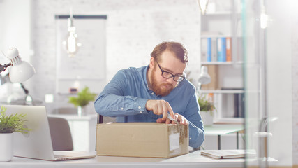 Shot of a Creative Man Opens Up Parcel While Sitting at His Desk in Bright and Modern Office.