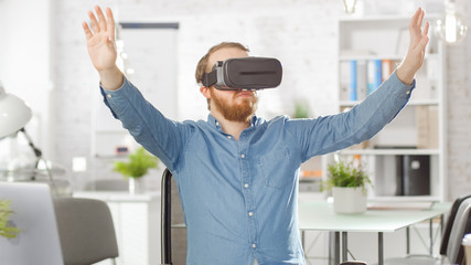 Naklejka na ściany i meble Creative Young Developer Uses Virtual Reality Glasses with Augmented Reality Technology while Sitting at His Desk. He is Working in a Bright Modern Loft Office.