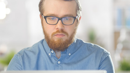 Fototapeta premium Portrait shot of a Bearded Creative Young Man Wearing Glasses Sitting in His Bright Office and Working on a Personal Computer. Computer Screen Reflects in His Glasses.