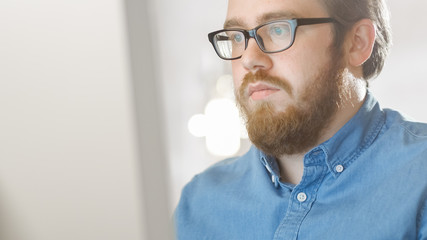 Portrait shot of a Bearded Creative Young Man Wearing Glasses Sitting in His Bright Office and Working on a Personal Computer. Computer Screen Reflects in His Glasses.