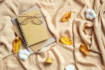 Autumn flat lay composition. Paper notebook, glasses, cotton and dry leaves on beige plaid. Autumn hygge style desk table