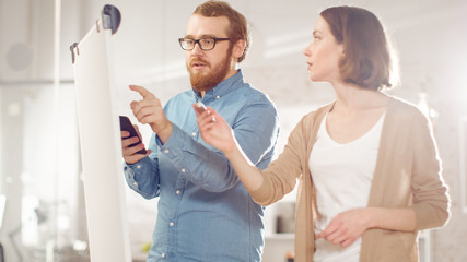 Obraz premium Young Man and a Woman Discuss Working Process on a Whiteboard. Man Holds Smartphone, woman Draws on a Whiteboard. Bright Modern High Tech Office with Beautiful People.