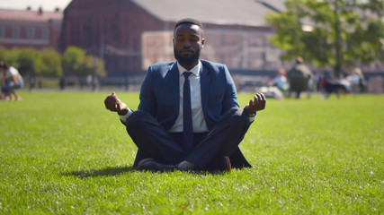 Relaxed african businessman sitting on lawn doing meditation in city park