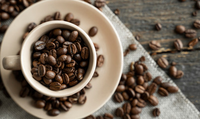Coffee cup filled of fresh arabica or robusta coffee beans with scattered coffee beans on a linen textile and wood table.