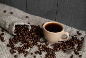 Coffee scattered from a linen bag and a cup of black coffee Espresso or Americano standing on a wooden table. Fresh arabica coffee beans on linen textile. Aromatic freshly brewed coffee.